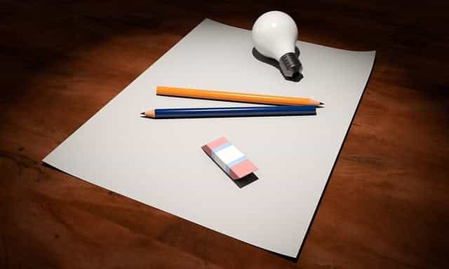 A blank sheet of paper lying flat on a wooden table with two pencils, an eraser and a lightbulb on top of it.