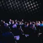 A group of people sit in the audience of a conference, watching a presentation.