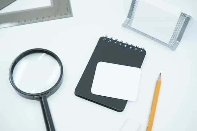 A magnifying glass, notepad and pencil sit on a white desktop.