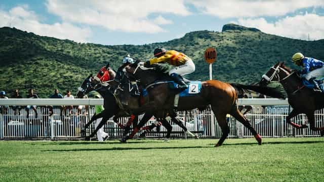 Four jockeys racing their horses at full speed on an outdoor track, with rolling green mountains and hills behind them.