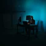 A masked and shadowy figure sitting at a desk in front of two computer monitors while being drenched in blue light.